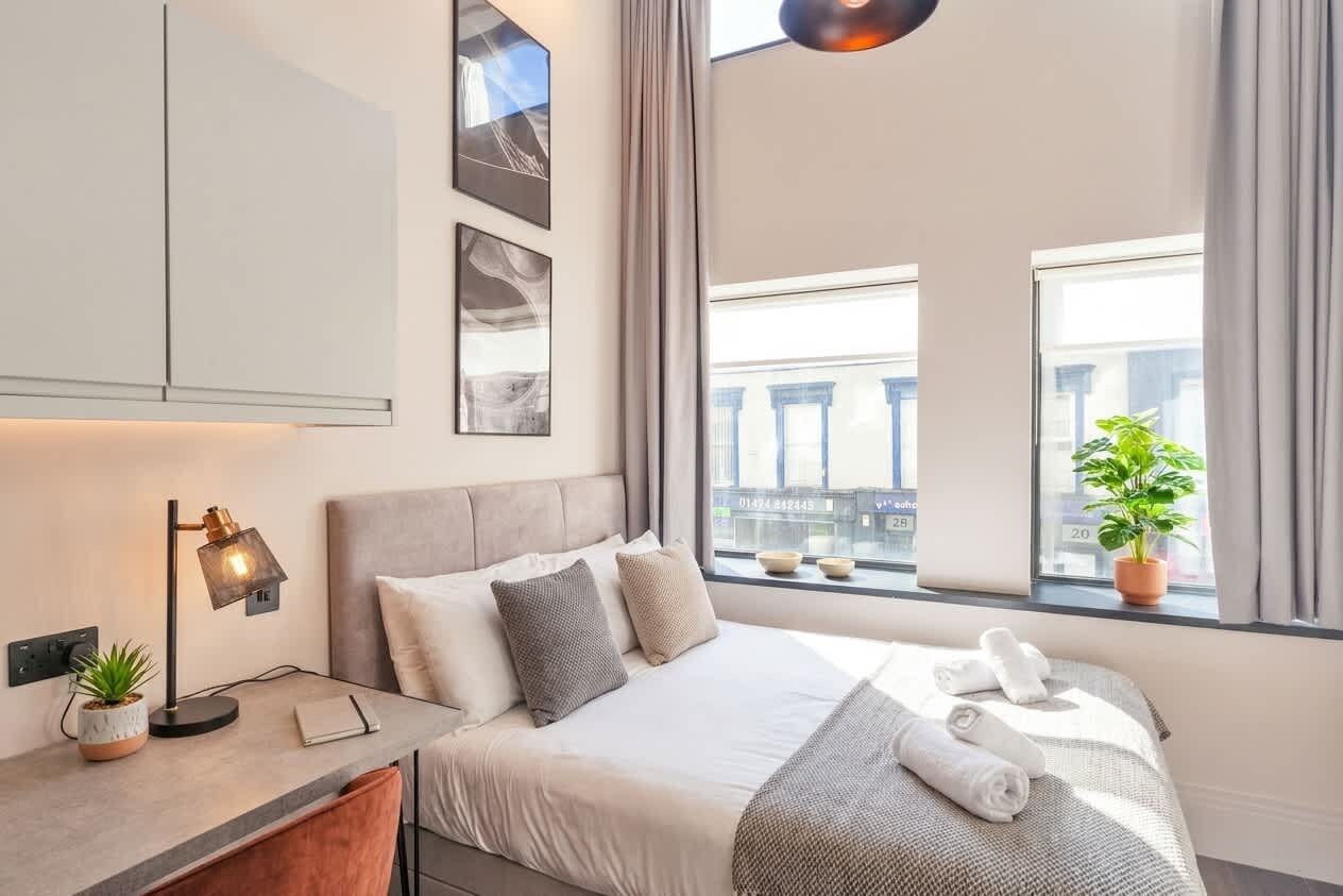 Habitació d'hotel moderna amb llit, taula de nit, làmpada i vista de la ciutat a través de grans finestres.
