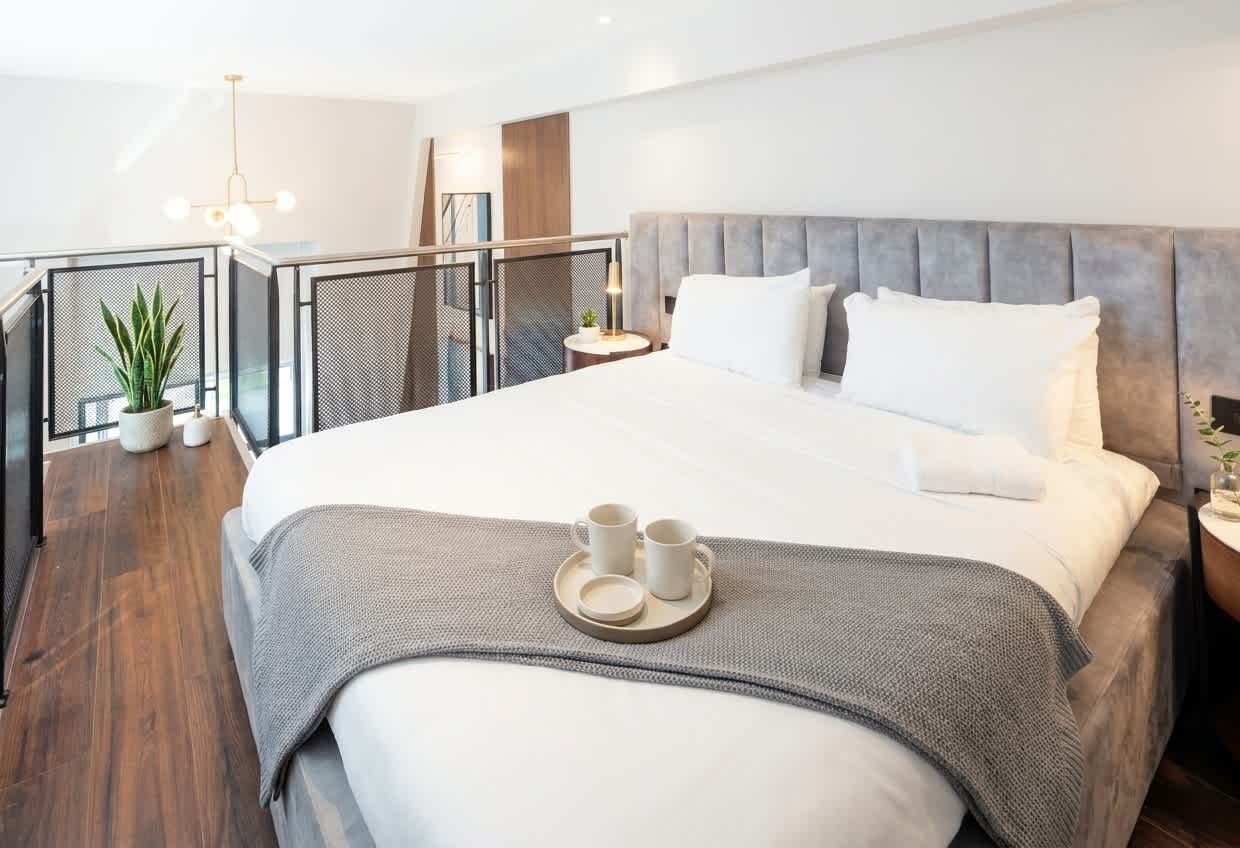 Una habitación de hotel moderna con una cama grande, una mesita de noche pequeña, una planta en maceta y un balcón con barandilla.