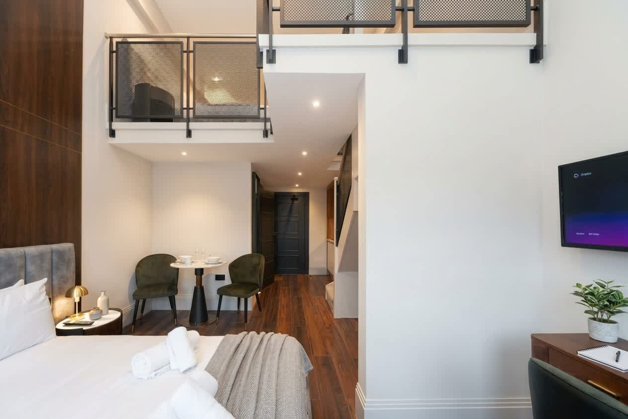 Et moderne hotelværelse med en hævet balkon, en seng med pænt sengetøj, et skrivebord med stol og et fjernsyn på væggen.