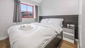 A neatly made bed with white linens, a bedside table with a power outlet, and a window with red frames and grey curtains. - Budget Double Studio Room in Central Nottingham (Nottingham)