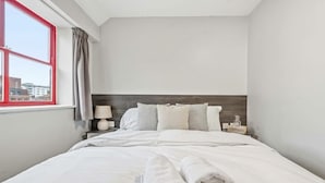 A hotel room with a bed, a nightstand, a lamp, a window with curtains, and a view of buildings outside. - Cosy Budget Double Studio in Nottingham Centre (Nottingham)