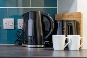 Un coin cuisine comprenant une bouilloire électrique noire, deux tasses blanches et une planche à découper en bois.