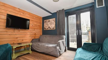 A room with a television mounted on a wooden wall, a grey sofa, a small green table, and a sliding glass door leading outside.