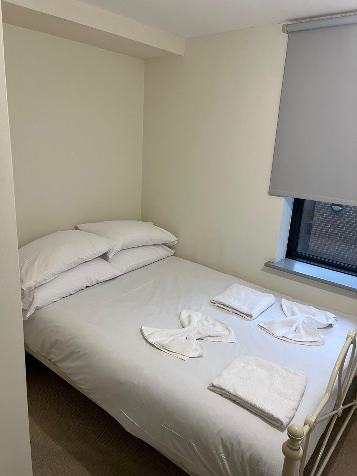 A single bed with white linens, two pillows, and a window with a blind.
