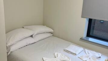 A single bed with white linens, two pillows, and a window with a blind.