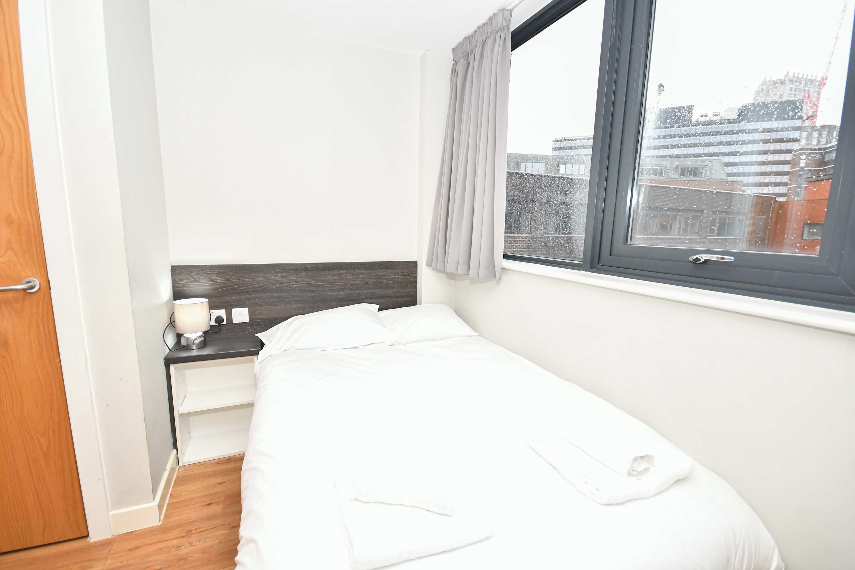 Une petite chambre d’hôtel, bien rangée, avec un lit, une table de chevet, une lampe et une fenêtre avec des rideaux.