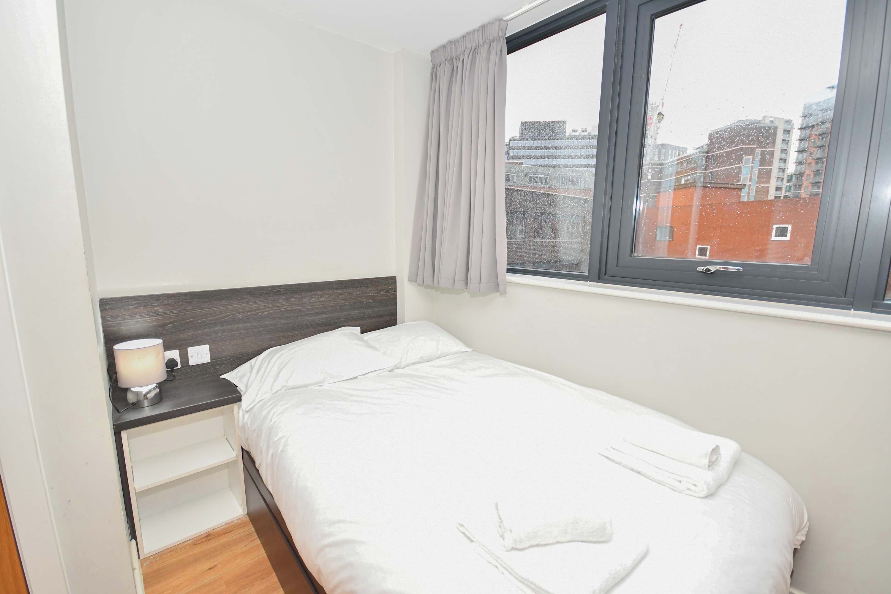 Una cama individual con ropa blanca, una mesita de noche con lámpara, una ventana con vistas a edificios y una cortina gris.