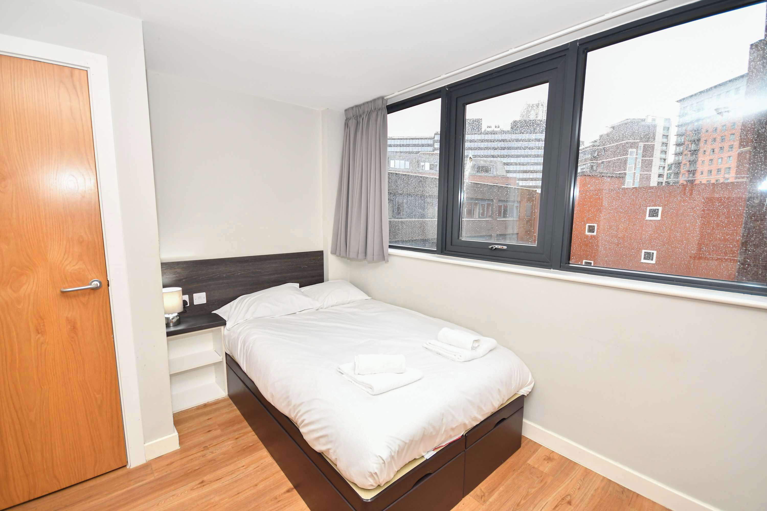 A bedroom with a bed, a nightstand, a window with a view of buildings, and a wooden door.