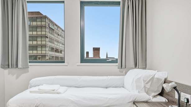 A hotel room with a bed, a desk with a lamp, two windows with curtains, and a view of a building outside. - Modern Budget Single Studio in Central Nottingham (Nottingham)
