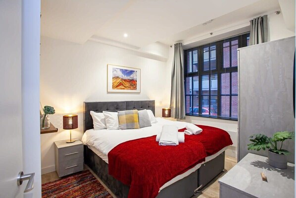 A modern hotel room with a large bed, a bedside table with a lamp, a window with blinds, and a painting on the wall. - Contemporary 2 Bed Apartment in Central Manchester (Manchester)