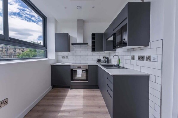 A modern kitchen with dark cabinetry, a stainless steel oven, a microwave, and a window with a view of buildings. - Modern and Bright Studio in Central Sheffield (Sheffield)