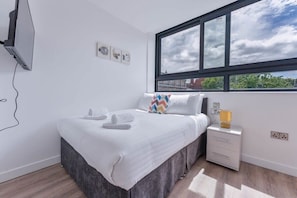 Ein Schlafzimmer mit Bett, Fenster, Nachttisch und an der Wand montiertem Fernseher.
