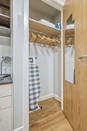 A walk-in closet with wooden shelves, hanging wooden hangers, a checkered ironing board, and a mirror on the door. - Central Nottingham Cosy Budget Double Studio (Nottingham)