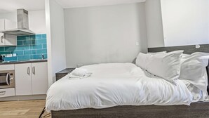 Un dormitorio con cama, ropa de cama blanca, cabecera de madera, kitchenette con microondas y salpicadero de azulejos azules.