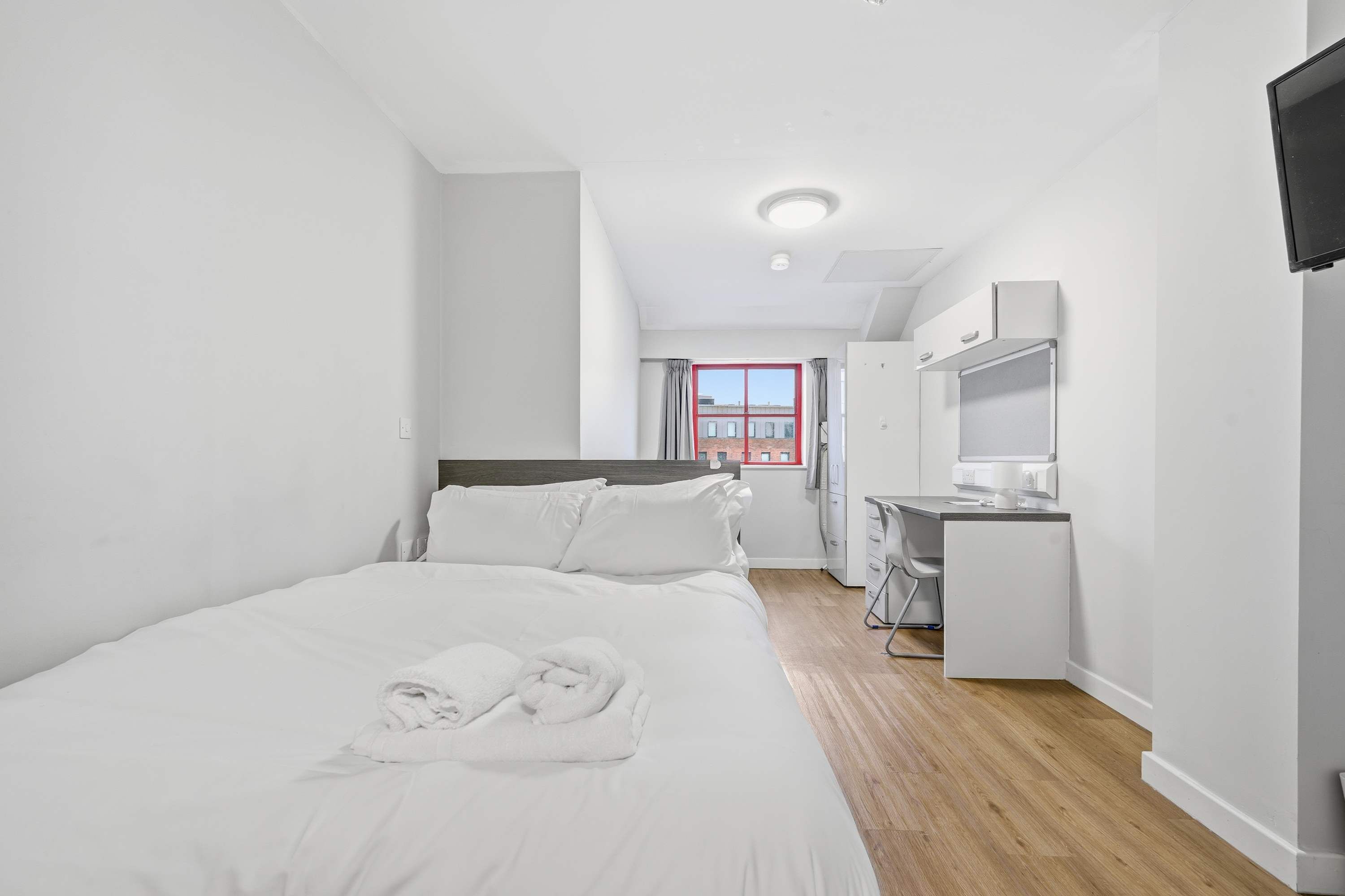 A modern hotel room with a bed, a desk, a chair, a TV, and a window with a red frame.