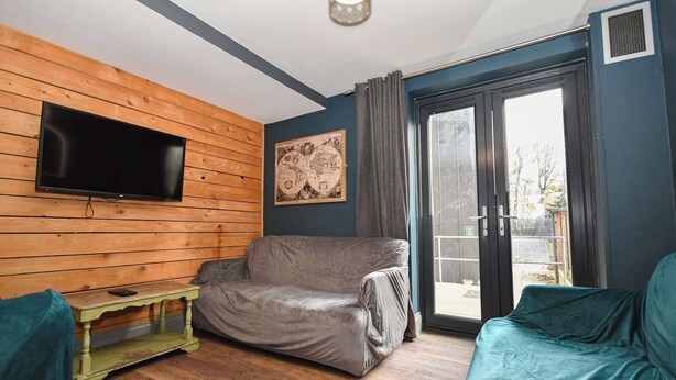 Ein Zimmer mit Holzwand, einem flachbildfernseher über einem grünen Tisch, einem grauen Sofa und einer Glastür zum Außenbereich.
