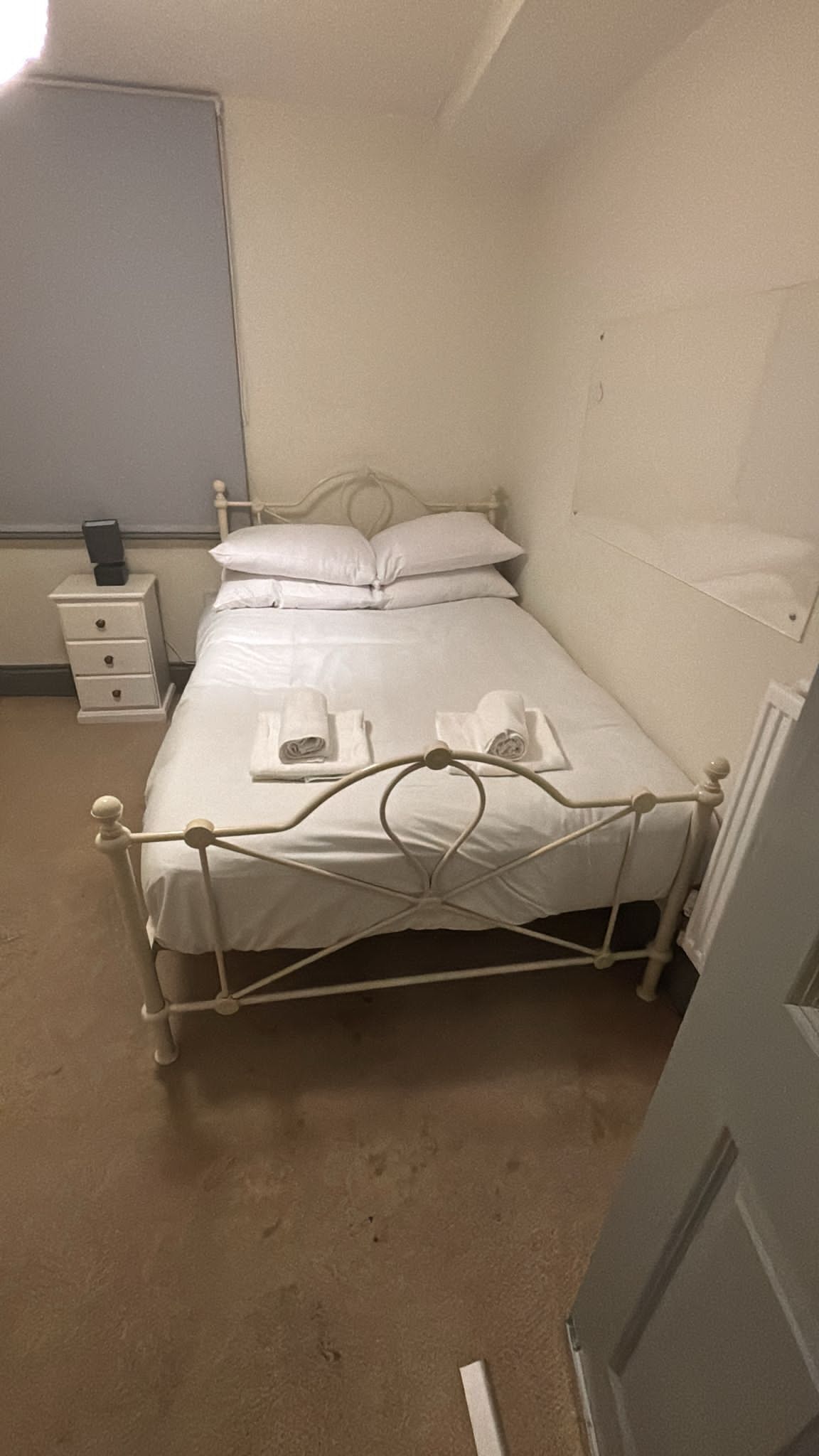 Ett litet rum med en enkel säng, en sängram i metall, vita sängkläder och ett nattduksbord med en bok på.