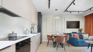 A modern kitchen with integrated appliances, a dining area with a round table and chairs, and a living room with a sofa and a TV. - Family Apartment with Balcony | Air Conditioning, Parking | Tarczyński Arena (Wrocław)