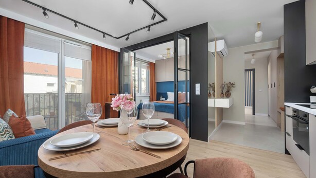 A modern dining area with a round wooden table, chairs, and a view of the outdoor balcony. - Family Apartment with Balcony | Air Conditioning, Parking | Tarczyński Arena (Wrocław)
