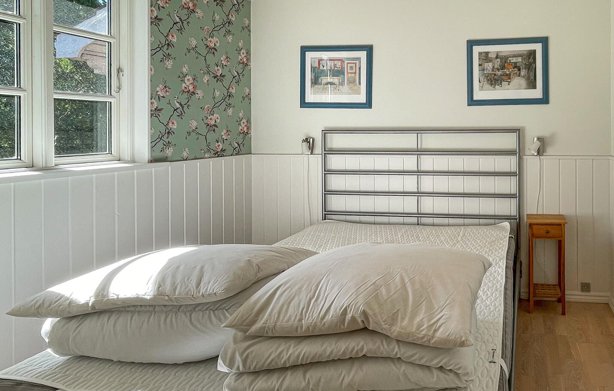 A bed with a metal headboard, a window with floral wallpaper, and two framed pictures on the wall.
