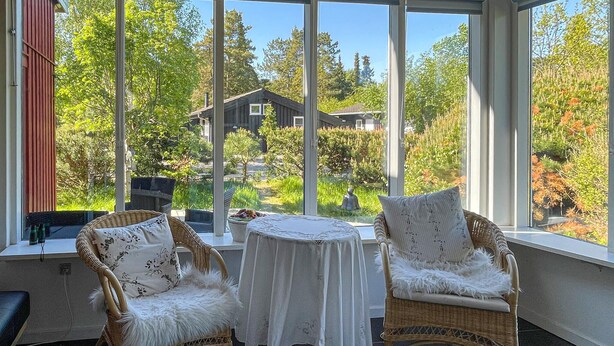 Ett soligt rum med flätade stolar, ett litet bord och utsikt över ett hus genom stora fönster.