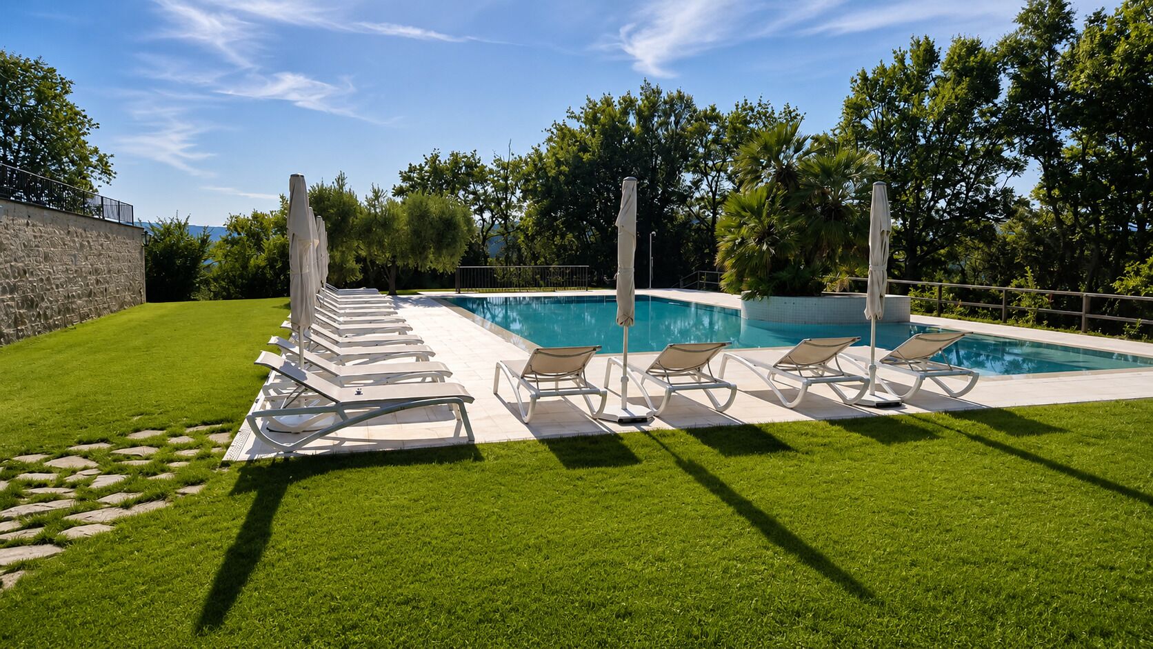 A swimming pool with lounge chairs and umbrellas, surrounded by a well-maintained lawn and trees.