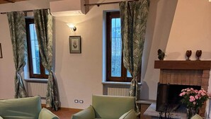 A living room with two green armchairs, a glass coffee table, a fireplace, and a window with curtains. - Cascina Lungamerenda - Relax in Monferrato (Costigliole d'Asti)