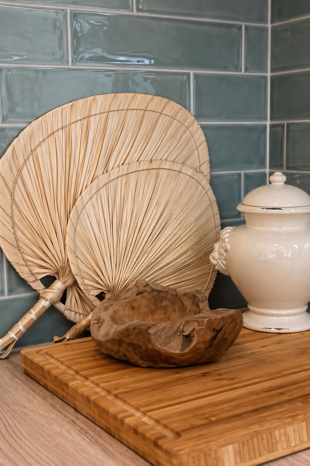 A wooden cutting board, a woven fan, and a ceramic pot on a kitchen counter.