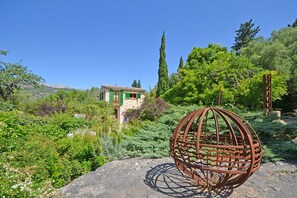 Ein rustikales Steinhaus mit grüner Dachdeckung, umgeben von üppiger Vegetation und einer Metallskulptur.