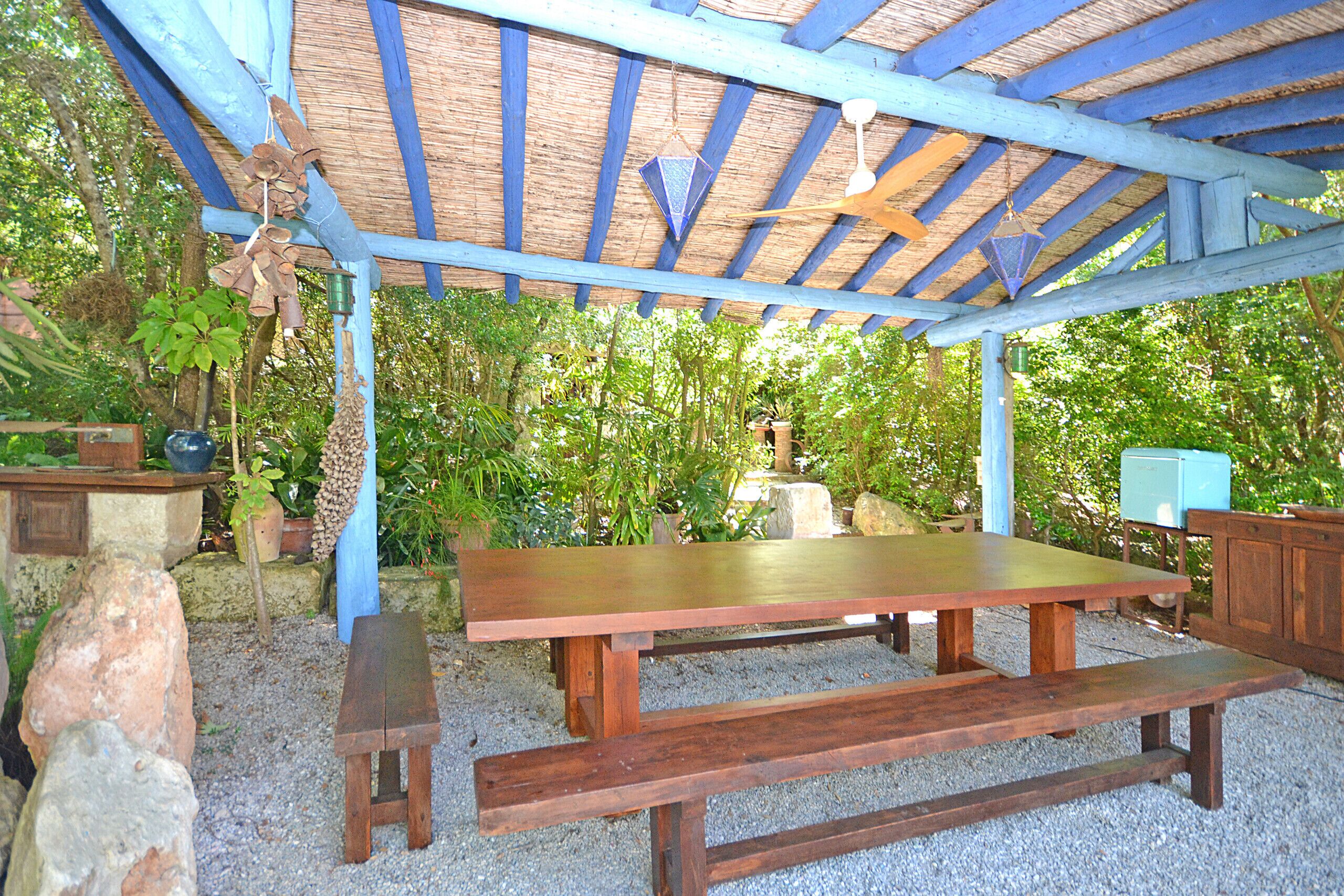 An outdoor wooden table and benches under a blue-roofed structure surrounded by greenery.