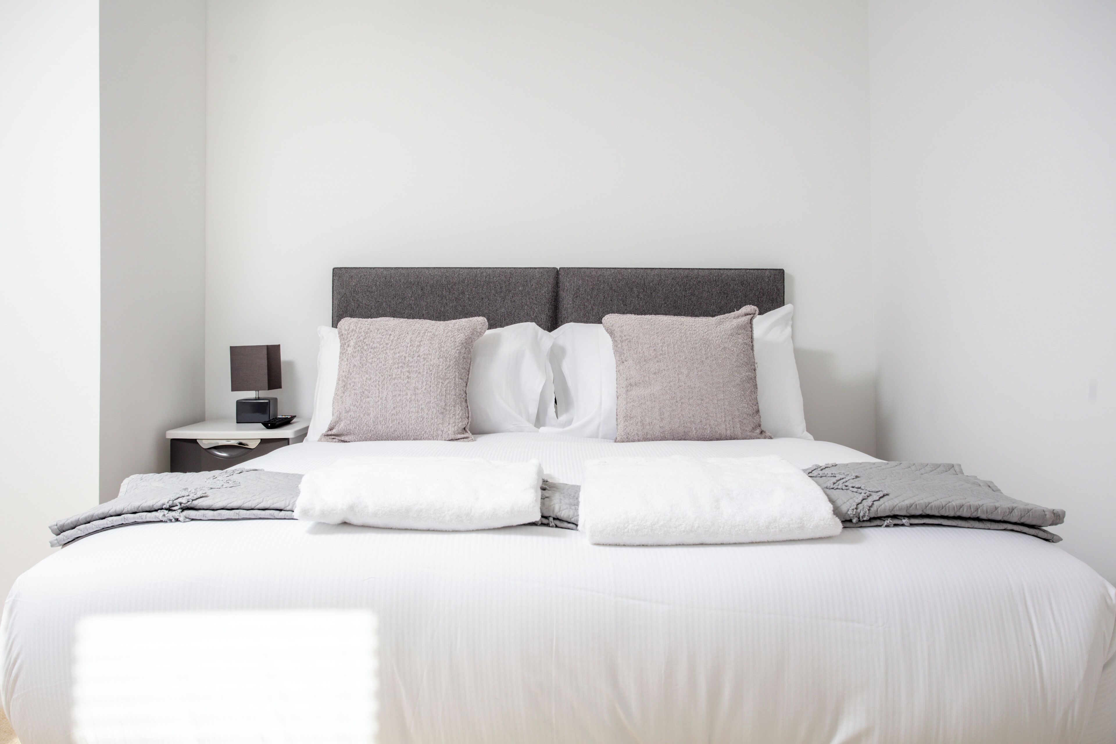 A neatly made bed with a grey headboard, white bedding, and a bedside table with a lamp.