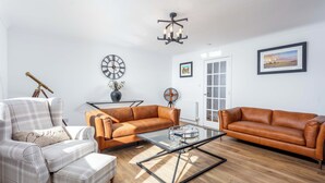 A living room with a brown leather sofa, a plaid armchair, a glass coffee table, a clock, and a fan. - The Lookout - Donnini Apartments (Prestwick)