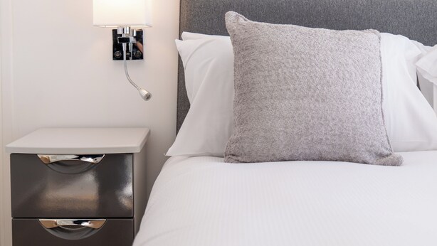 A neatly made bed with a grey headboard, white bedding, and a bedside lamp. - The Lookout - Donnini Apartments (Prestwick)