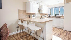 A modern kitchen with white cabinets, a wooden countertop, a microwave, and a coffee maker. There are three white bar stools and a window with a view. - The Lookout - Donnini Apartments (Prestwick)
