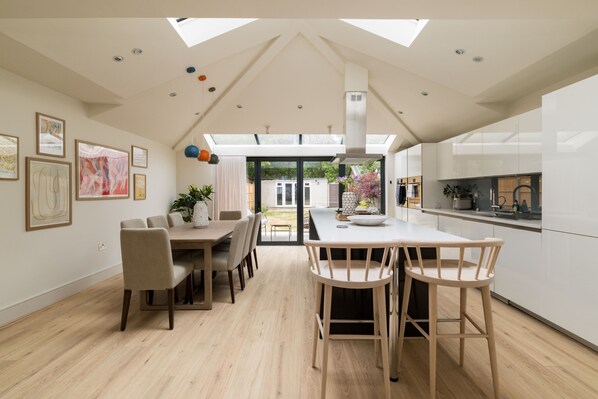 A modern kitchen with a central island, dining area with chairs, and a large window overlooking a garden. - The Glazed Gable & Alfresco (London)