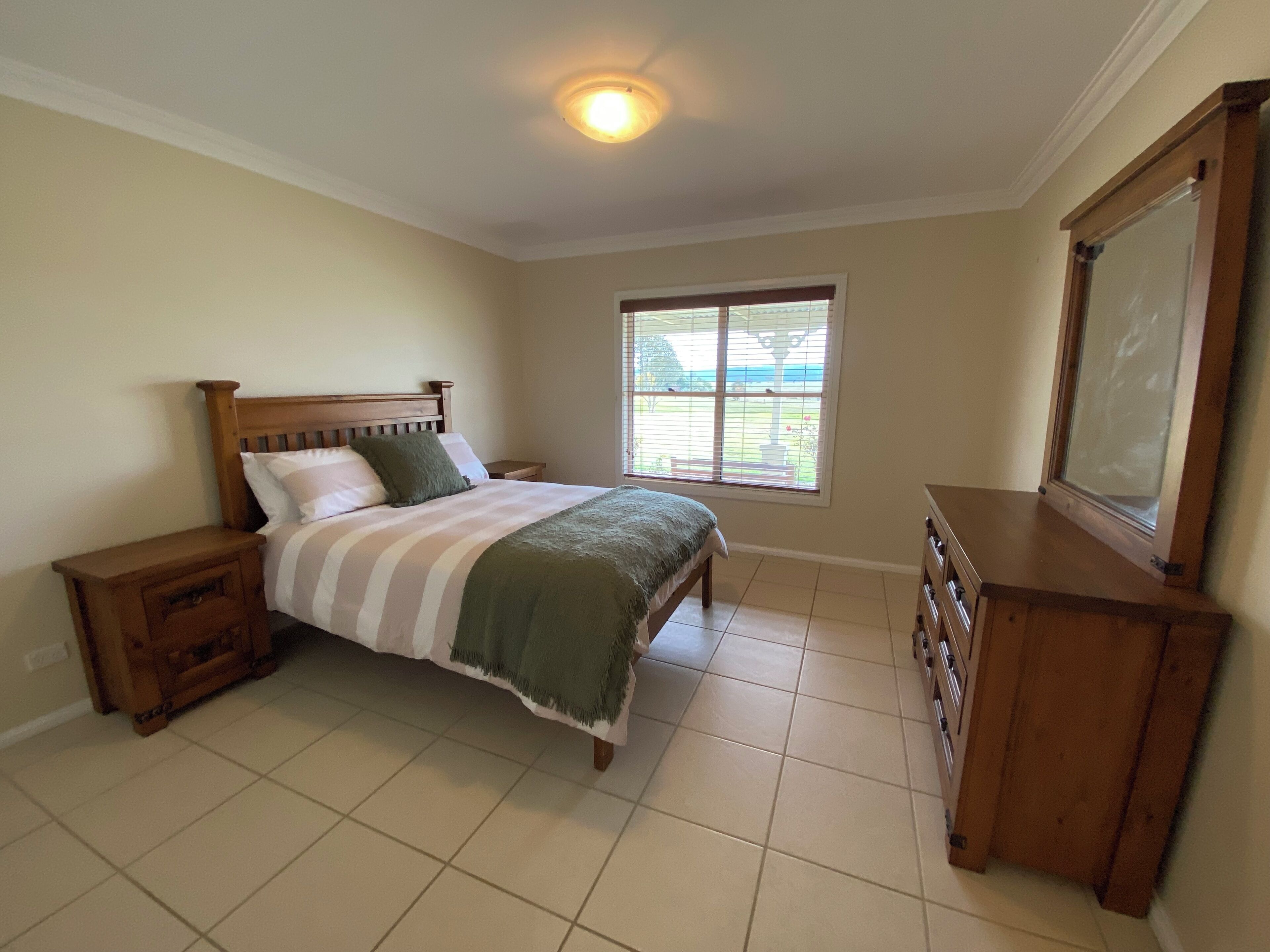 Une chambre à coucher comprenant un lit, des tables de chevet, une commode, une fenêtre avec des stores et un luminaire au plafond.