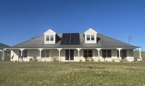 A two-story house with a large solar panel on the roof, a covered porch with benches, and a grassy yard.