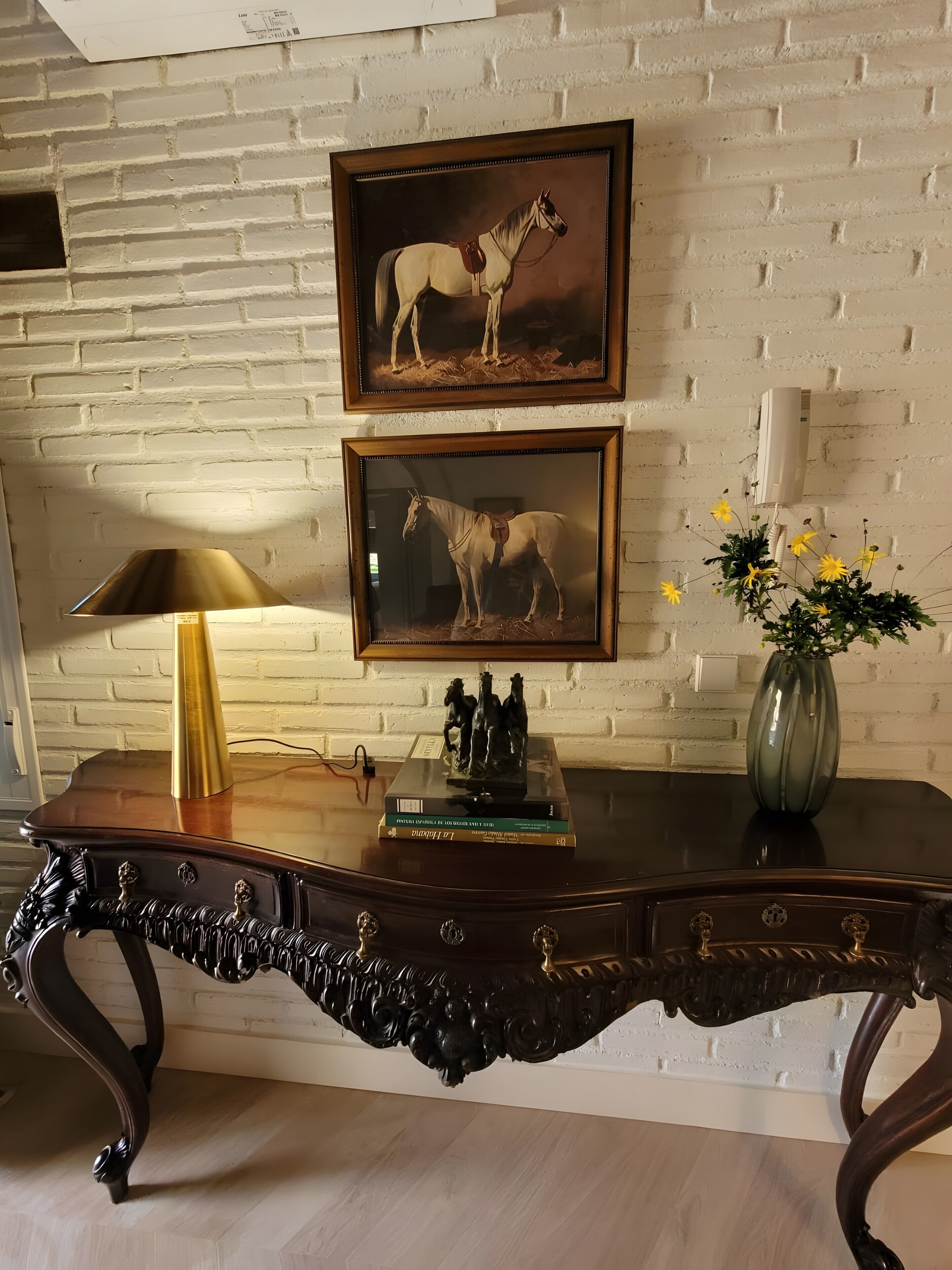 A wooden desk with ornate carvings, two framed horse paintings, a lamp, a vase with flowers, and a small statue on the desk.