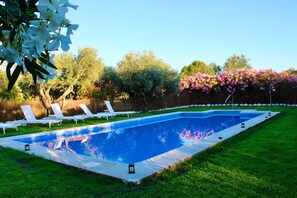 Una piscina rectangular con borde de azulejos, rodeada por un césped y sillas de descanso blancas.