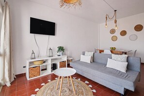 Ein Wohnzimmer mit grauem Sofa, weißen Kissen, rundem weißen Tisch, einem an der Wand befestigten Fernseher und einer Deckenleuchte.