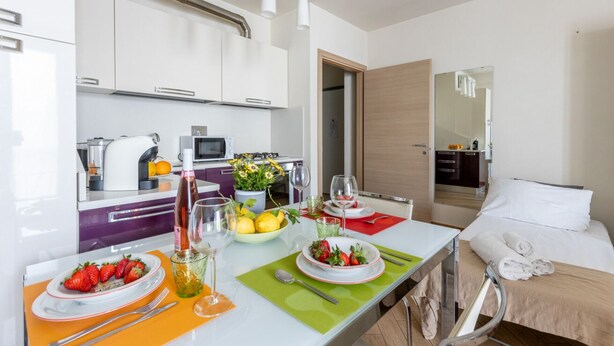Una habitación de hotel moderna con kitchenette, una zona de comedor con una mesa preparada para dos personas y una cama con toallas.
