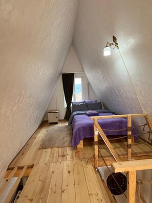 Une petite chambre mansardée de forme triangulaire, avec un lit, un banc en bois et un luminaire suspendu.