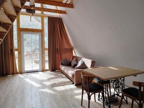 Ein Zimmer mit Holzbalkendecke, einer Couch mit Kissen, einem Esstisch mit StĂŒhlen und einer GlastĂŒr zum Garten.