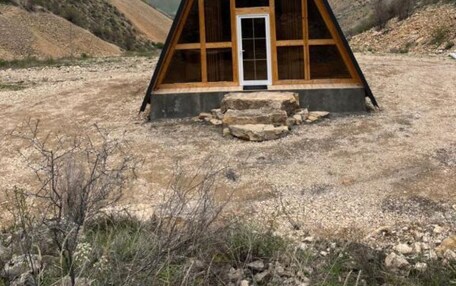 A triangular wooden structure with a central door and a small window, surrounded by a dry, rocky landscape with mountains in the background.