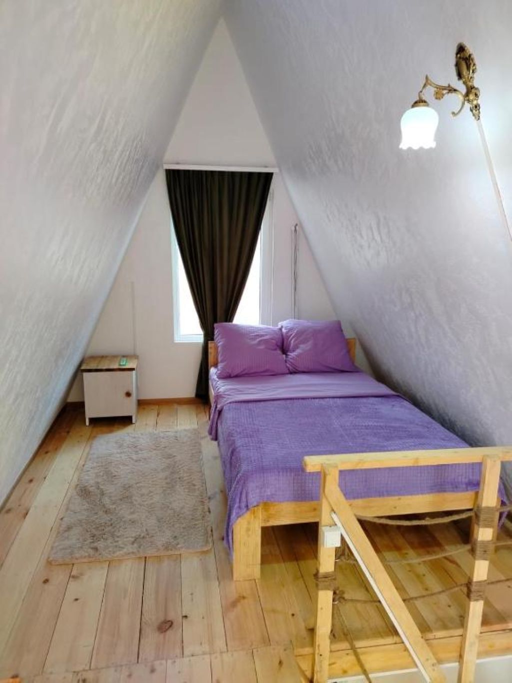 Un dormitorio con techo inclinado, cama con ropa de cama púrpura, una mesita de noche pequeña y un banco de madera.