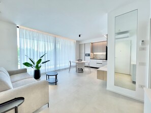 Ein modernes Wohnzimmer mit einer Couch, einem Esstisch mit Stühlen, einer Küchenzeile mit einer Mikrowelle und einem großen Fenster mit durchsichtigen Vorhängen.