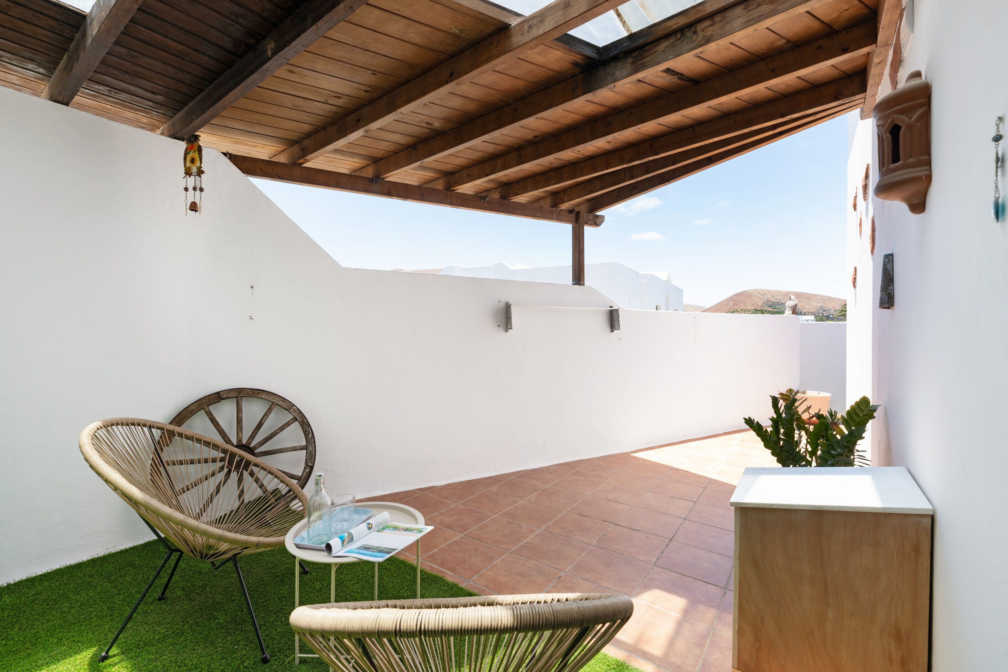 A covered patio with a wooden ceiling, a wicker chair, a small table with a bottle and a menu, and a potted plant.