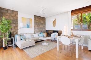 A modern living room with a stone wall, a fireplace, a dining area with white furniture, and a large window with plants.