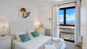 Een slaapkamer met een bed, nachtkastjes, een raam met gordijnen, een radiator en uitzicht op zee.