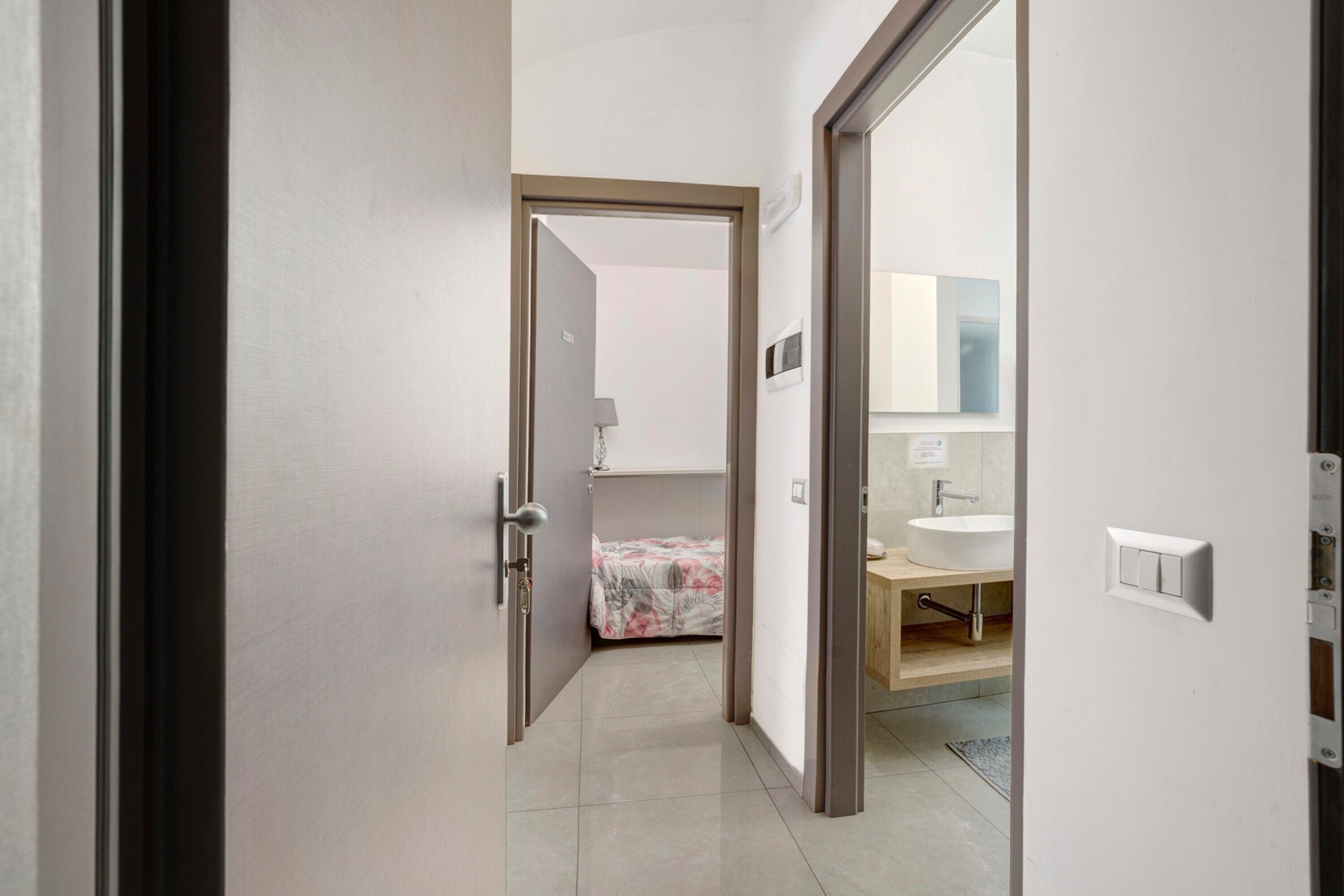 Un corridoio conduce a una camera da letto con un letto, un bagno con un lavandino e uno specchio.